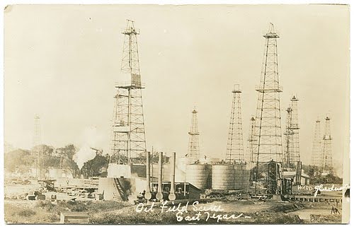Black-and-white photo of an early oil field in East Texas, featuring multiple tall oil derricks, storage tanks, and various industrial equipment scattered across the site. The image is labeled “Oil Field Scene East Texas.”.